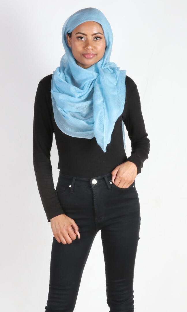 A beautiful 6ft tall female model in a profile pose displays a handmade cashmere shawl in aquamarine blue wrapped around her head and draped nicely around her neck to convey the message of how the cashmere shawl can be used in a different environments when circumstances call for it.