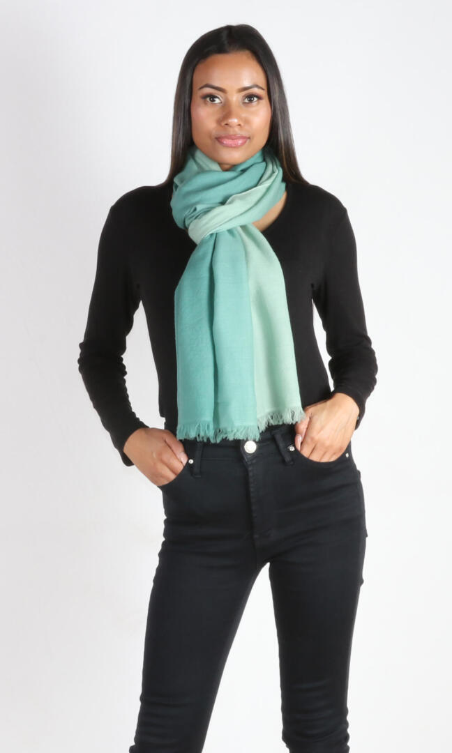 A beautiful 6ft tall female model in a profile pose displays a handmade cashmere shawl in sea green ombre color wrapped around her neck in a scarf knot to convey the message of supersoft and weightlessness of the shawl that can be used as a scarf in a wedding event.