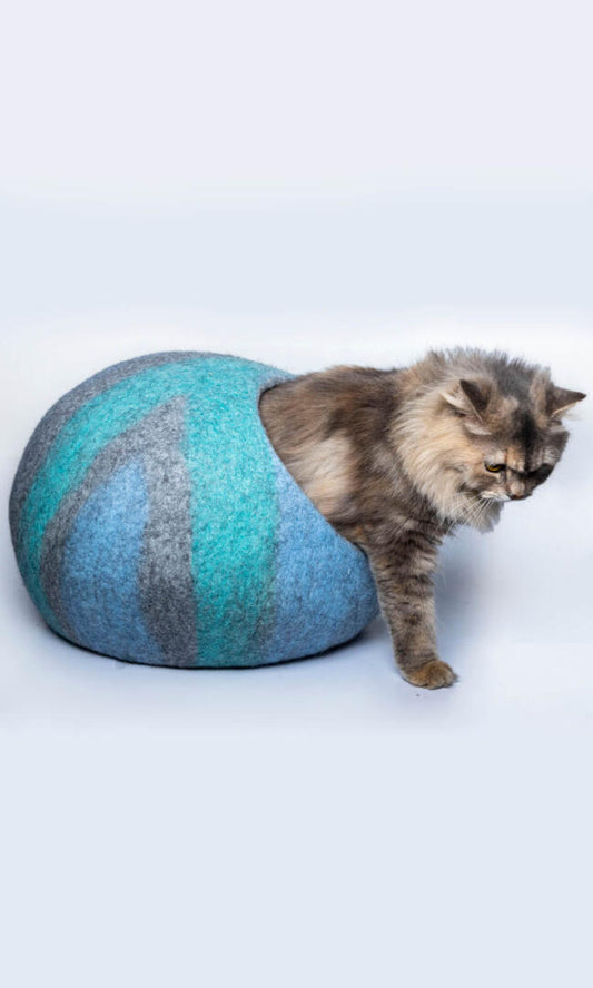 A cute cat comes out of its Abstract Oceanic Cat cave house with an oval-shaped bed. The cat house features a gray base with a sky blue & tiffany blue abstract paint-like design.