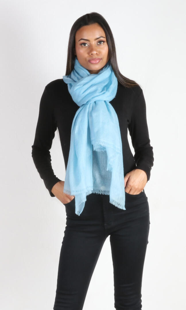 A beautiful 6ft tall female model in a profile pose displays a handmade cashmere shawl in aquamarine blue wrapped around her neck in a scarf knot to convey the message of supersoft and weightlessness of the shawl that can be used as a scarf.