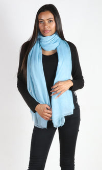 A beautiful 6ft tall female model in a profile pose displays a handmade cashmere shawl in aquamarine blue wrapped around her neck in a single loop to convey the message of the usability and versatility of the cashmere.