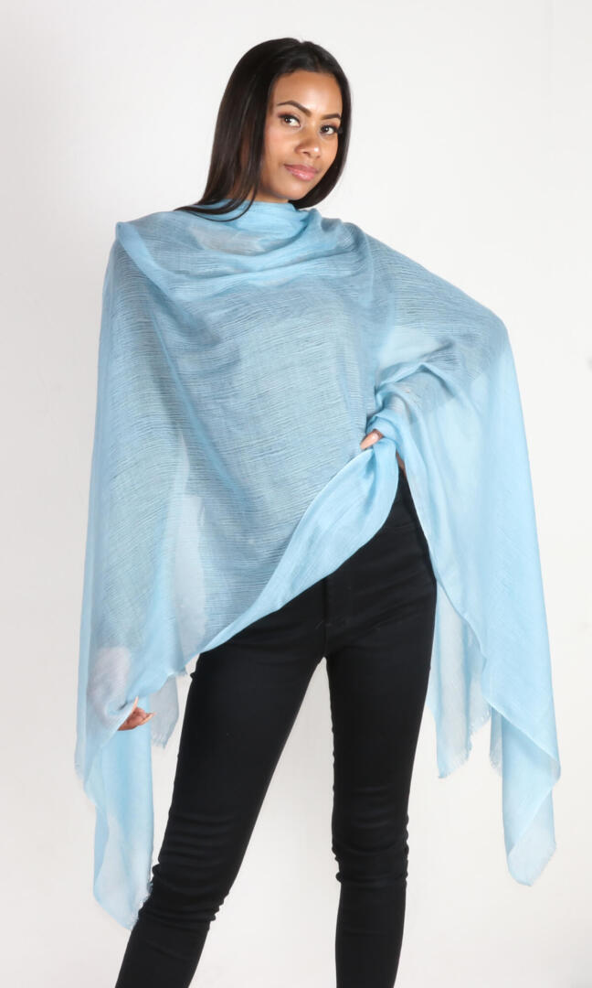 A beautiful female model in a profile pose displays a handmade cashmere shawl in aquamarine blue draped around her shoulders to convey the message of the elegance and breathability the shawl carries.