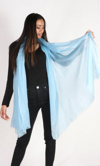 A beautiful female model displays a handmade cashmere shawl in aquamarine blue draped around her neck and she is holding the end of the shawl to convey the message that the shawl is lightweight and breathable.
