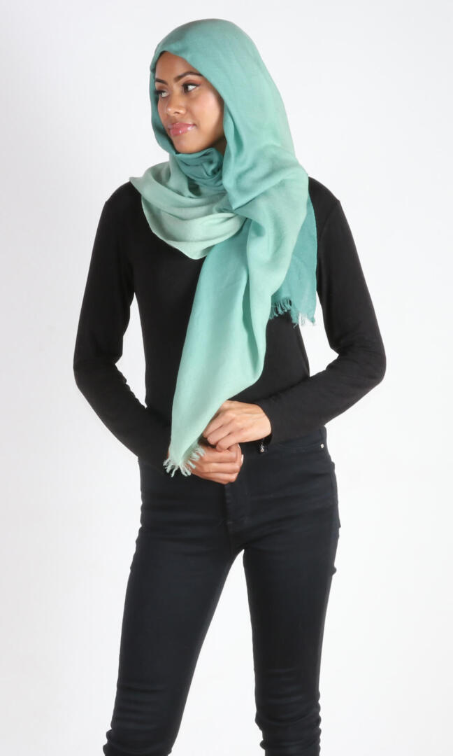 A beautiful 6ft tall female model in a profile pose displays a handmade cashmere shawl in sea green ombre color wrapped around her head and draped nicely around her neck to convey the message of how the cashmere shawl can be used in different environments when circumstances call for it.