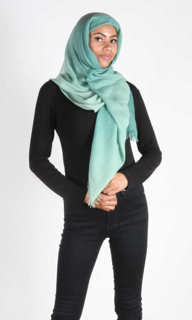 A beautiful 6ft tall female model in a profile pose displays a handmade cashmere shawl in sea green ombre color wrapped around her head as a hijab scarf to convey the message of the usefulness of the shawl in everyday life.