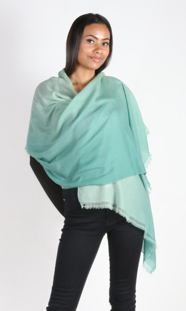 A beautiful 6ft tall female model in a profile pose displays a handmade cashmere shawl in sea green ombre color draped around her shoulders to convey the message of the elegance and breathability the shawl carries in a wedding event.