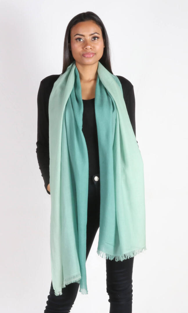 A beautiful 6ft tall female model in a profile view displays a handmade cashmere shawl in sea green ombre color draped from her neck to convey the message that the shawl has ample length to reach below the knees ideal for a wedding event or special day.