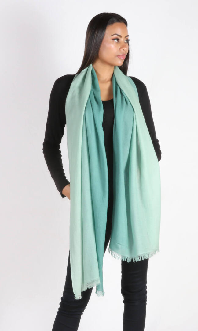 A beautiful 6ft tall female model in a profile view displays a handmade cashmere shawl in sea green ombre color draped from her neck to convey the message that the shawl has ample length to reach below the knees.